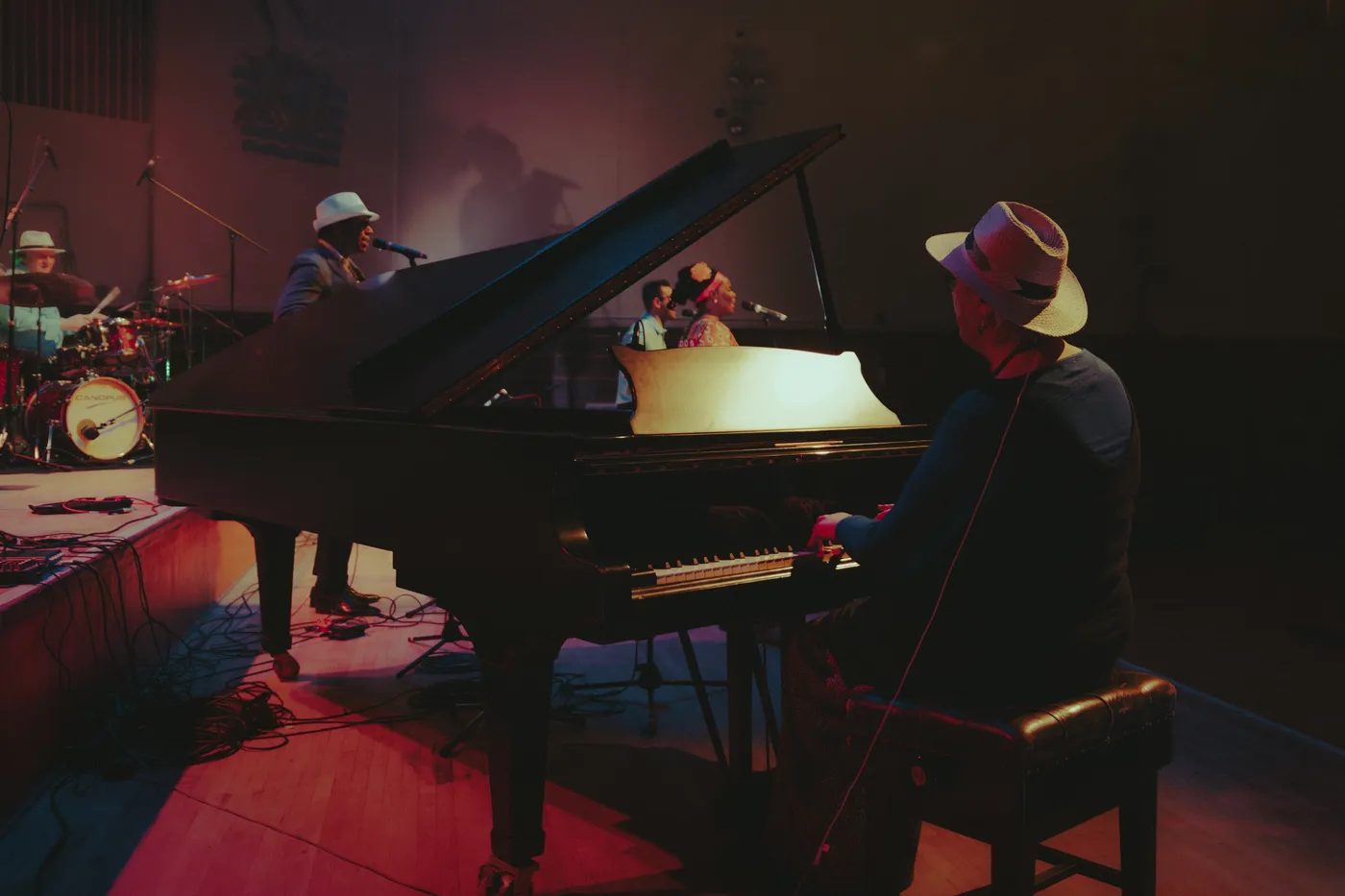 A pianist in a hat at a grand piano, with the drummer and other players in the background