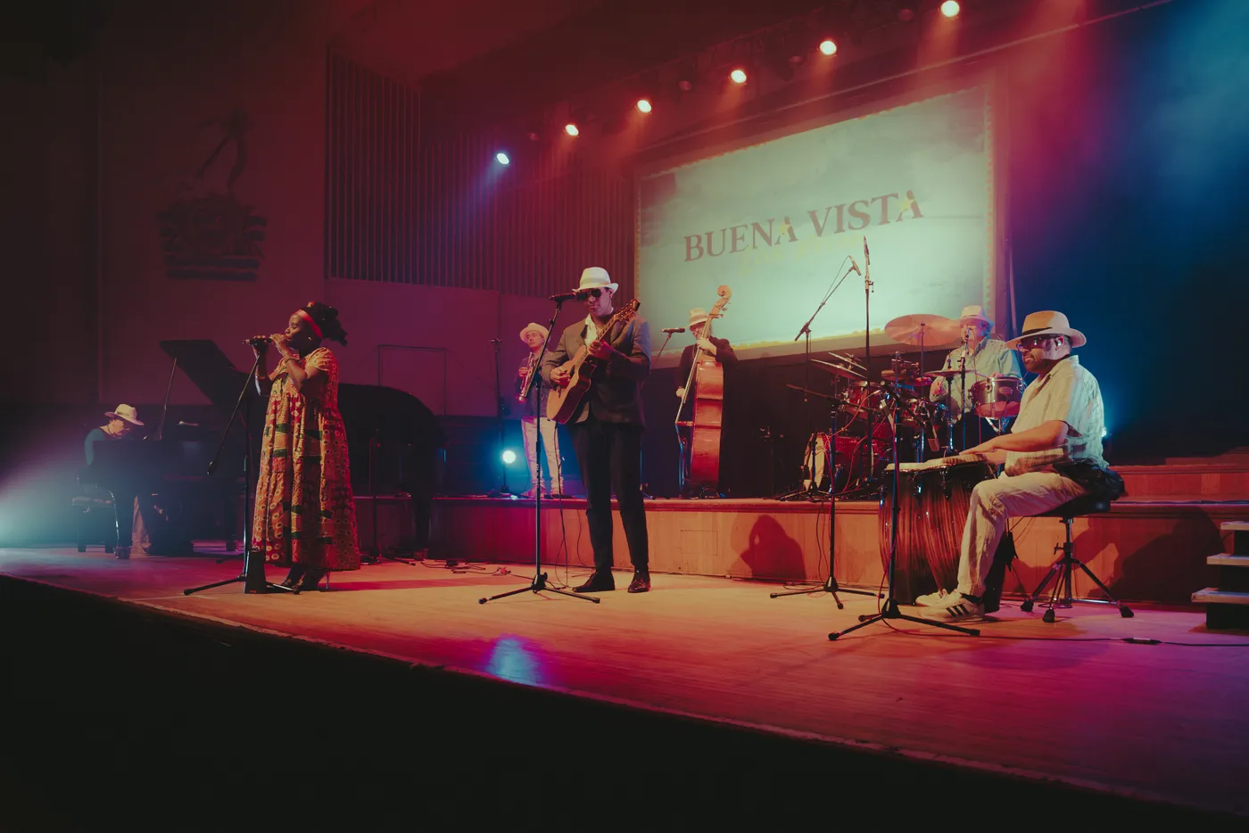 Buena Vista Live on a wide stage under red lights, a singer and guitarist at the front and a conga player on the right