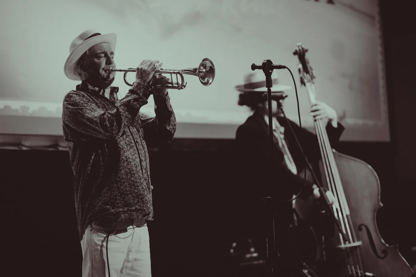 Black and white photograph of a trumpeter in a white hat playing, with a double bass behind him