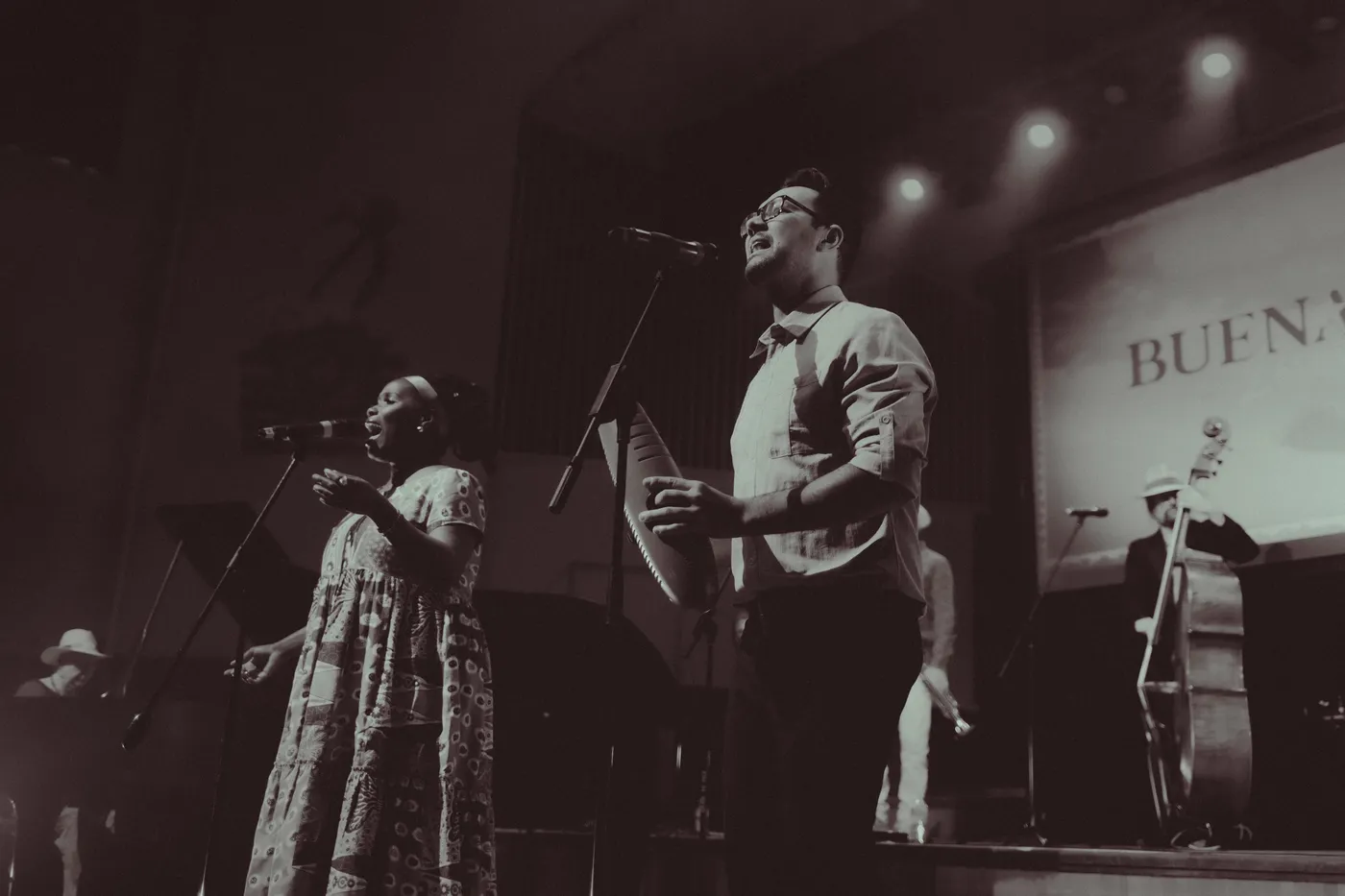 Black and white photograph of two singers at microphones on stage, with a double bass player at the back