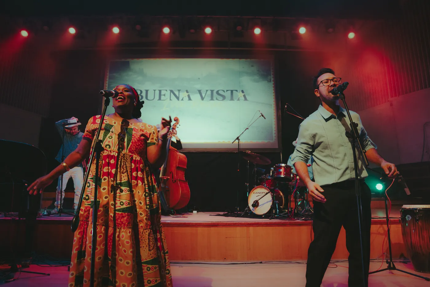 Two singers from Buena Vista Live performing at microphones in front of a screen reading Buena Vista, with the band behind them