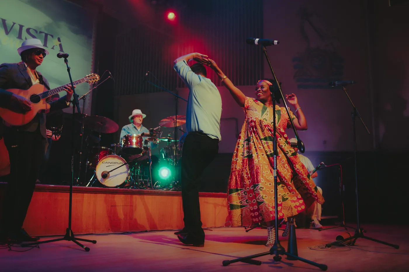 A singer in a patterned dress and a dancer in a pale shirt dancing at the front of the stage while a guitarist plays