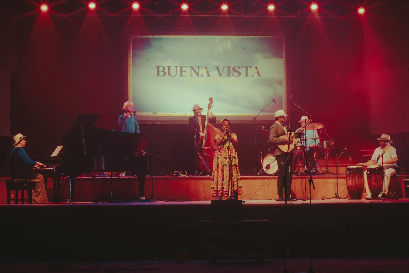 Wide view of the whole band on stage under red lights with the Buena Vista screen above