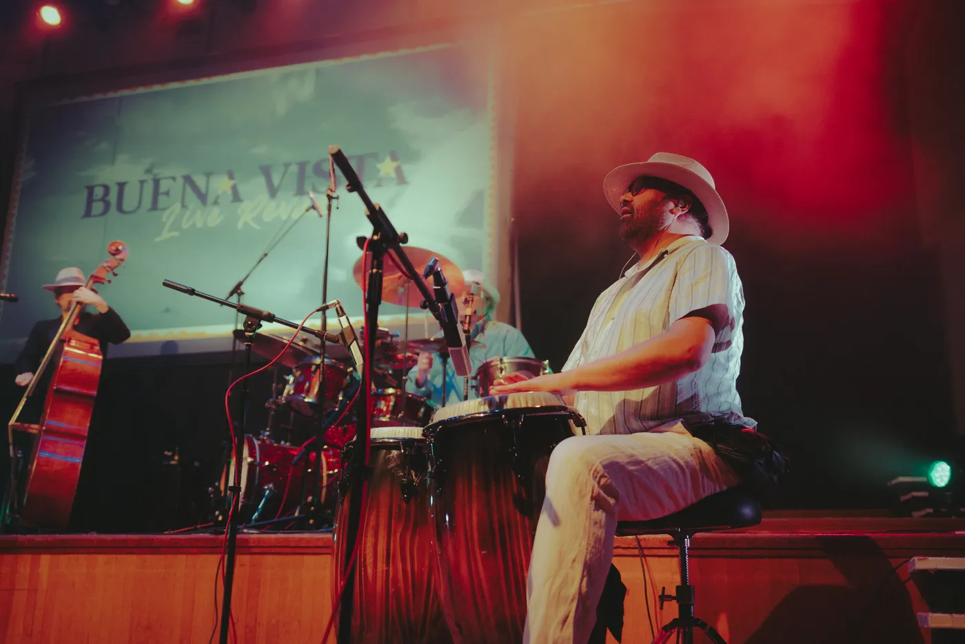 A conga player in a white hat playing at the front of the stage, with a double bass player behind