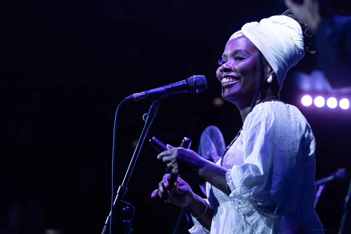 A singer in a white headwrap and white dress smiling at the microphone under blue stage light