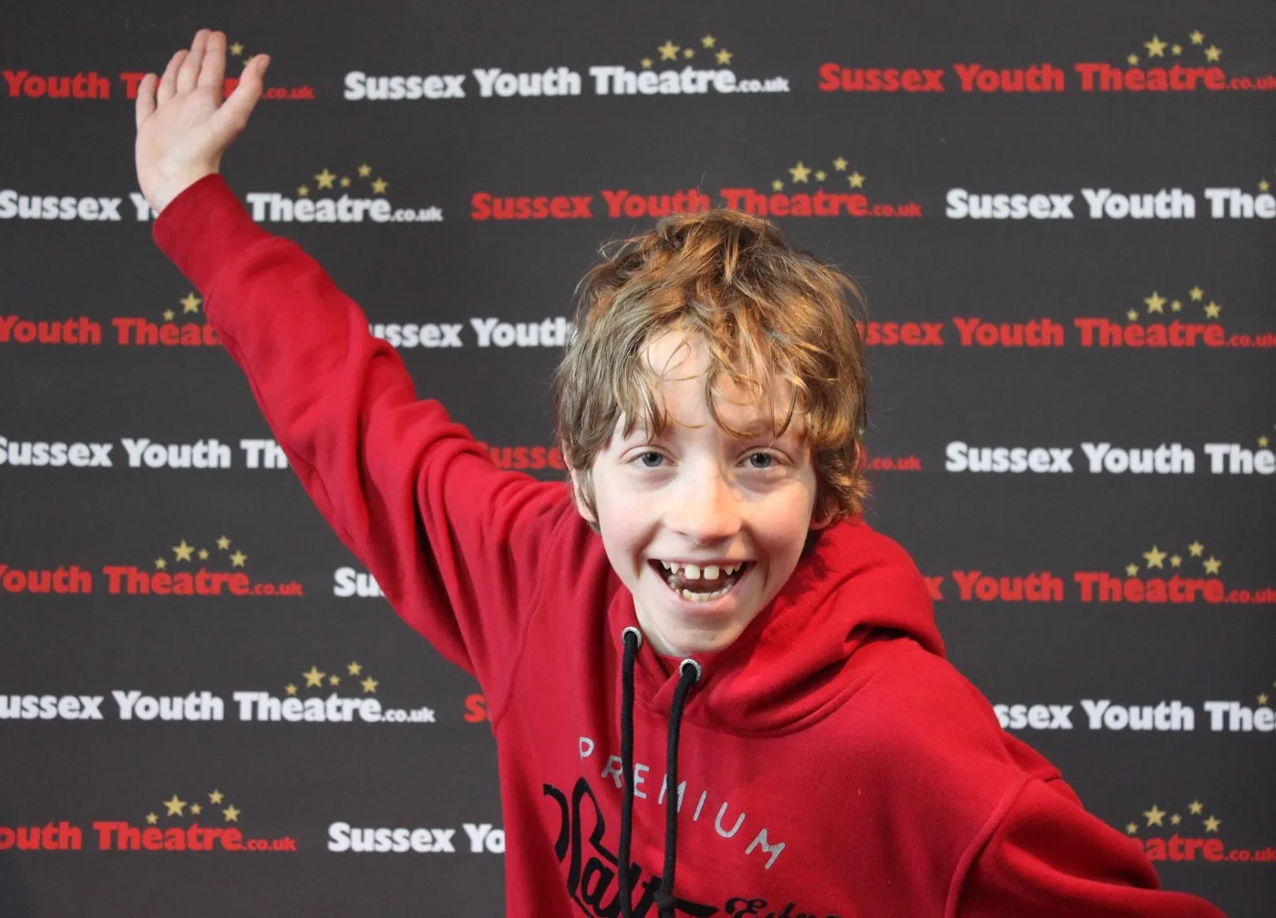 A smiling boy in a red hoodie with one arm raised, in front of a Sussex Youth Theatre branded backdrop