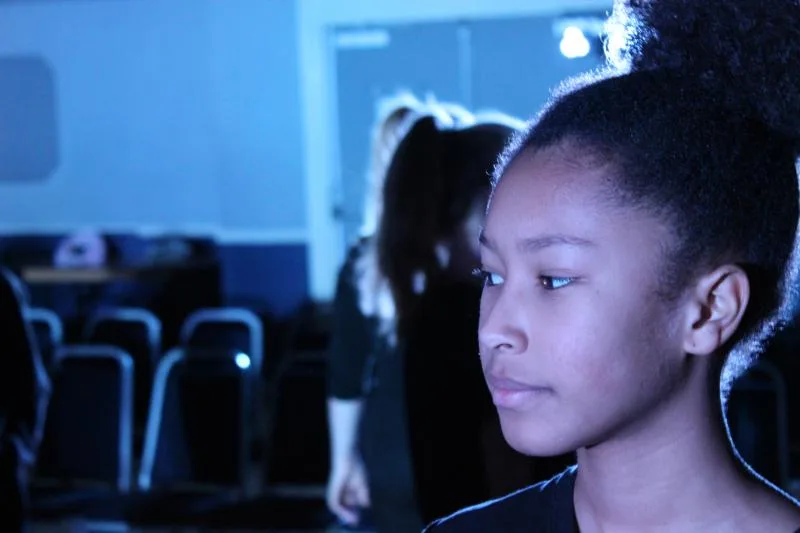 A girl in profile under blue stage lighting, with rows of chairs and other young people behind her