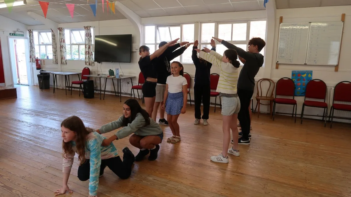 Young people in a hall with a wooden floor during a drama workshop, some standing with arms raised and some crouching on the floor