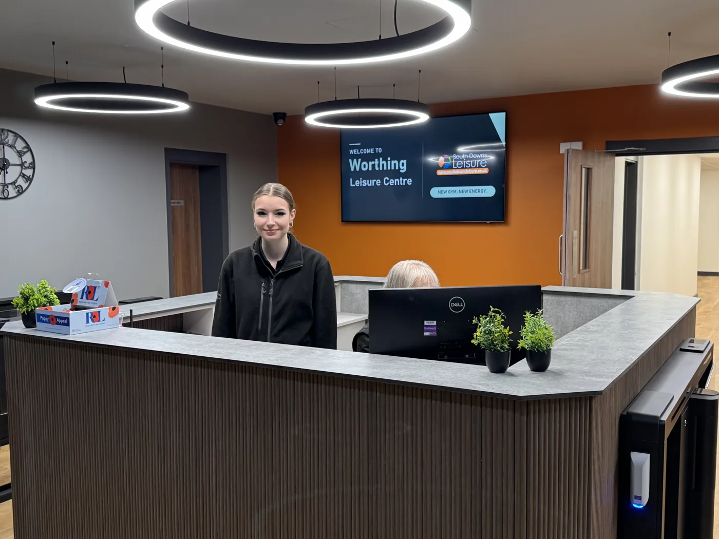 A member of staff standing behind the new reception desk at Worthing Leisure Centre, with a welcome screen on an orange wall behind