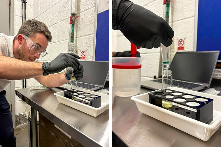 A man in safety glasses and black gloves pipettes a seawater sample into a row of small vials in a white tray at a steel lab bench